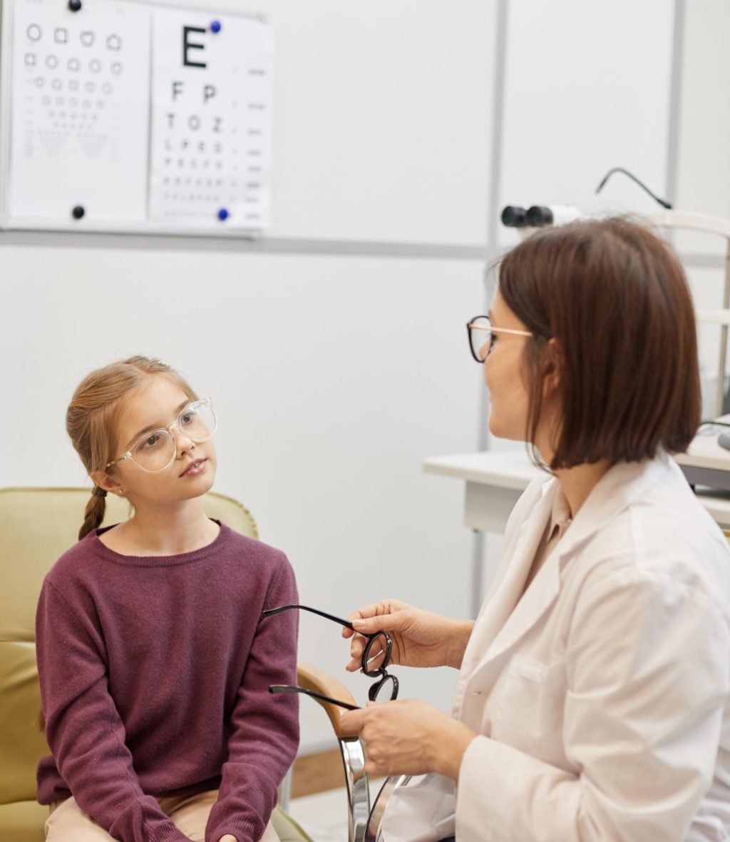 Portrait of cute girl trying on glasses during vision test in modern ophthalmology clinic, copy space